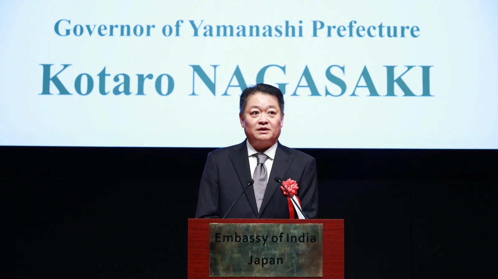 Yamanashi Governor Kotaro Nagasaki speaking at a podium marked Embassy of India, Japan.